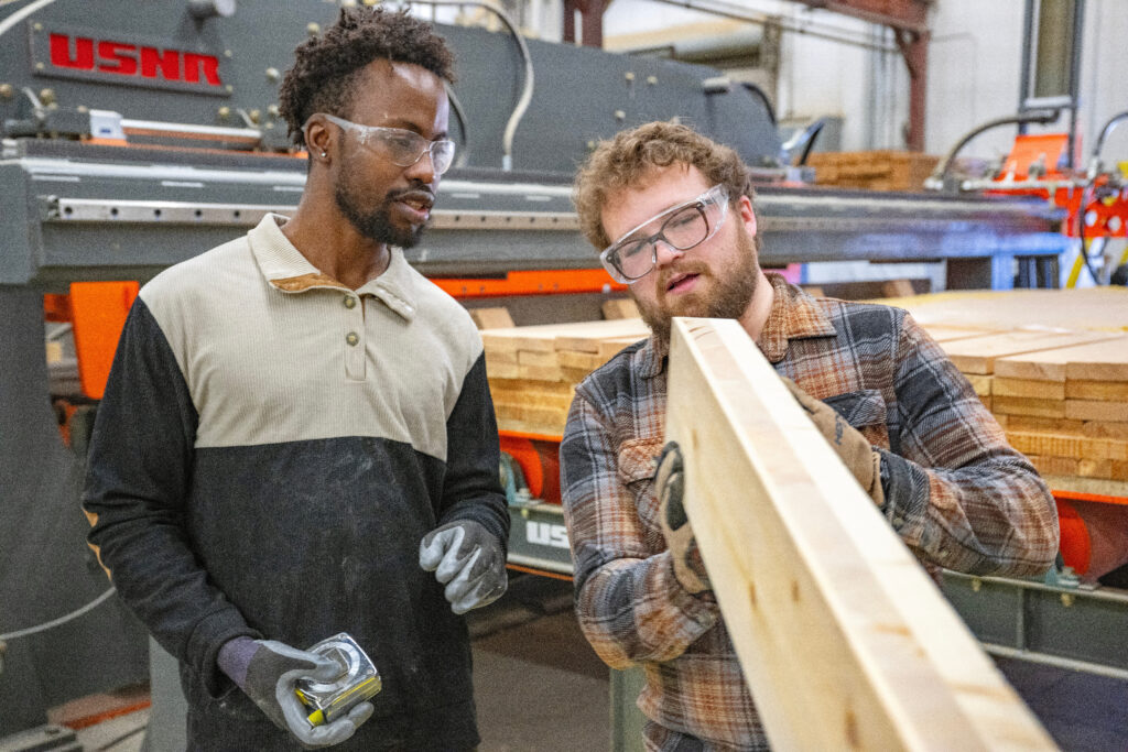 Two people wearing safety glasses examine a long wooden beam made of glued-together boards.
