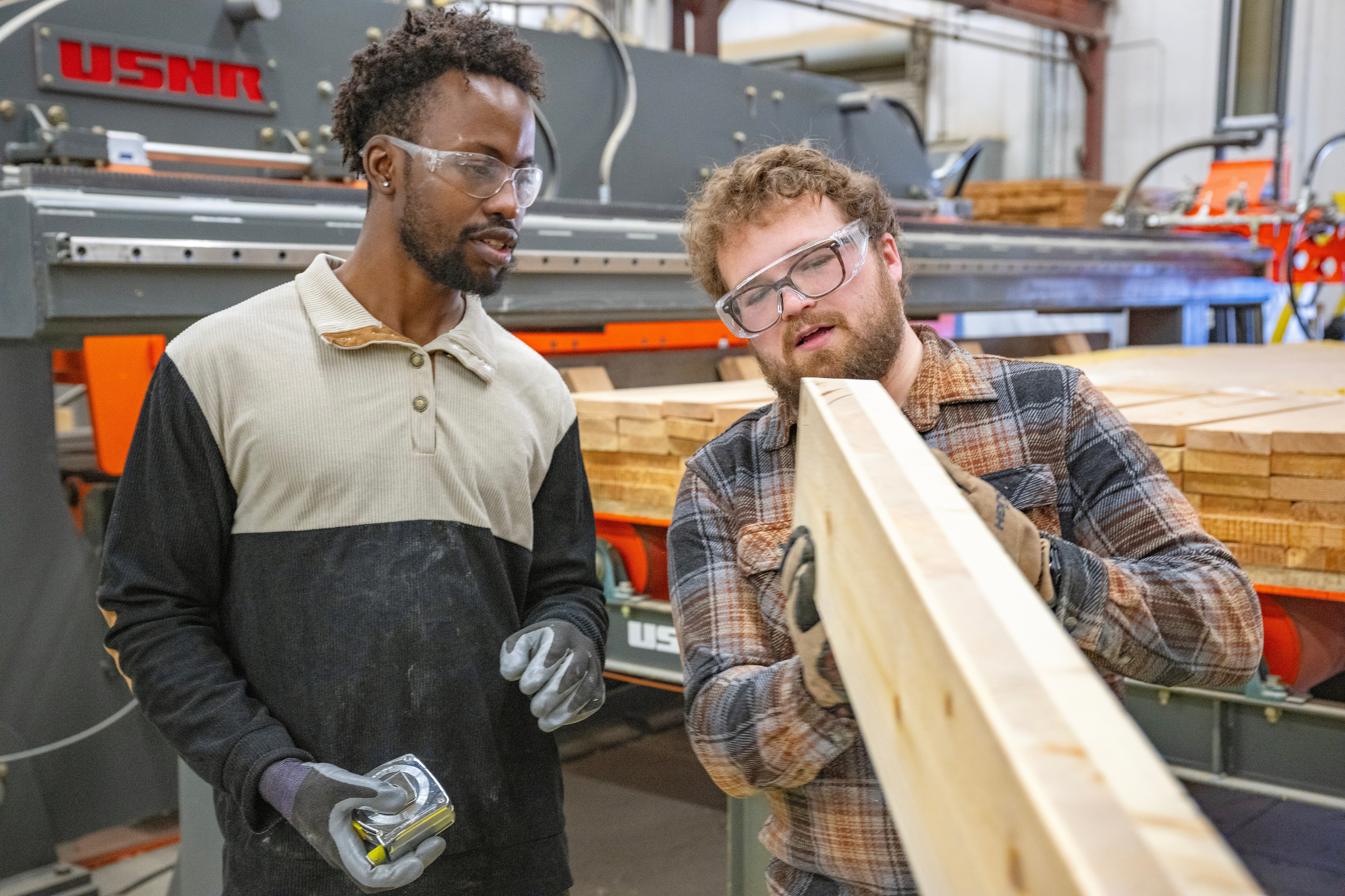 Two people wearing safety glasses examine a long wooden beam made of glued-together boards.