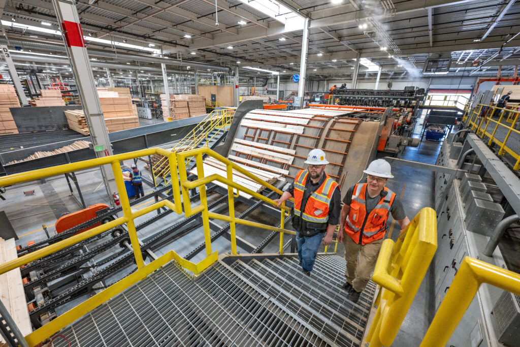 Two people in orange vests and hardhats talk in a massive industrial room with pallets of wood and a large piece of machinery in the background.