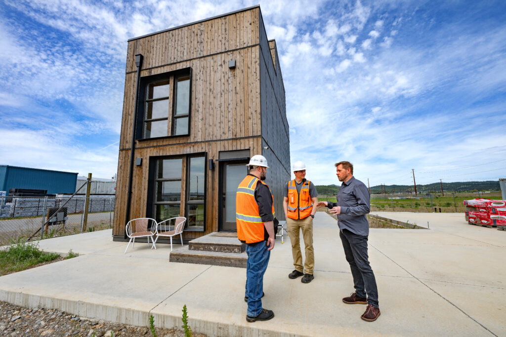 Three people, two in orange vests and hardhats, talk outside a small wood-paneled building made with mass timber.