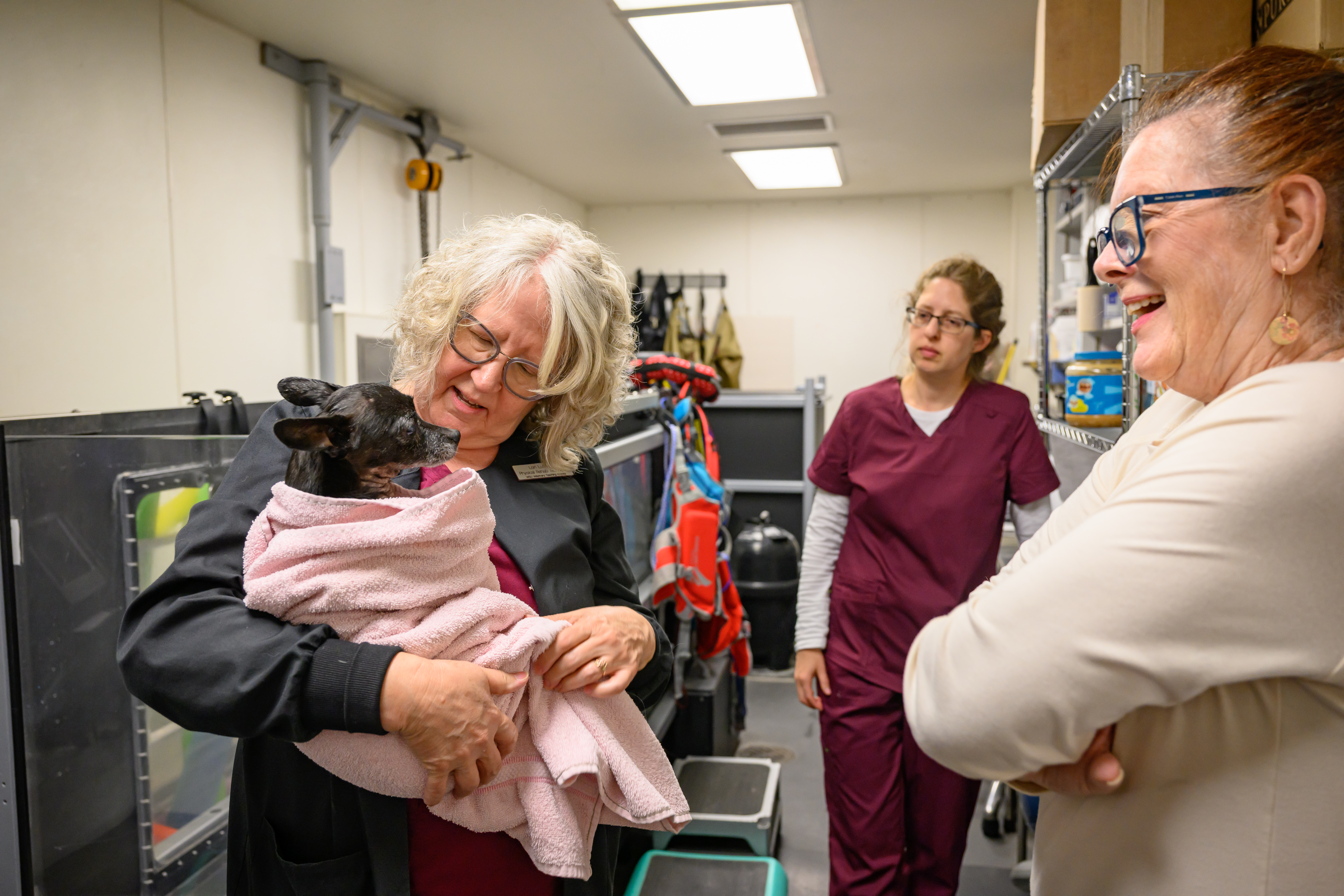 A person holding a small black dog wrapped in a towel while two other people look on.