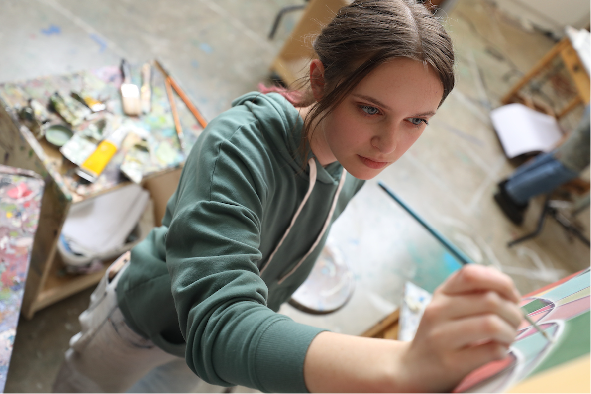 A person painting intently in an art studio with a table of paints and brushes behind them.