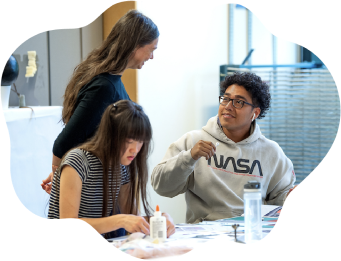 Two students seated at a table, one working and the other talking with a standing person.