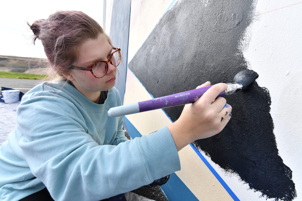 An artist wearing glasses painting with a brush on a large dark canvas