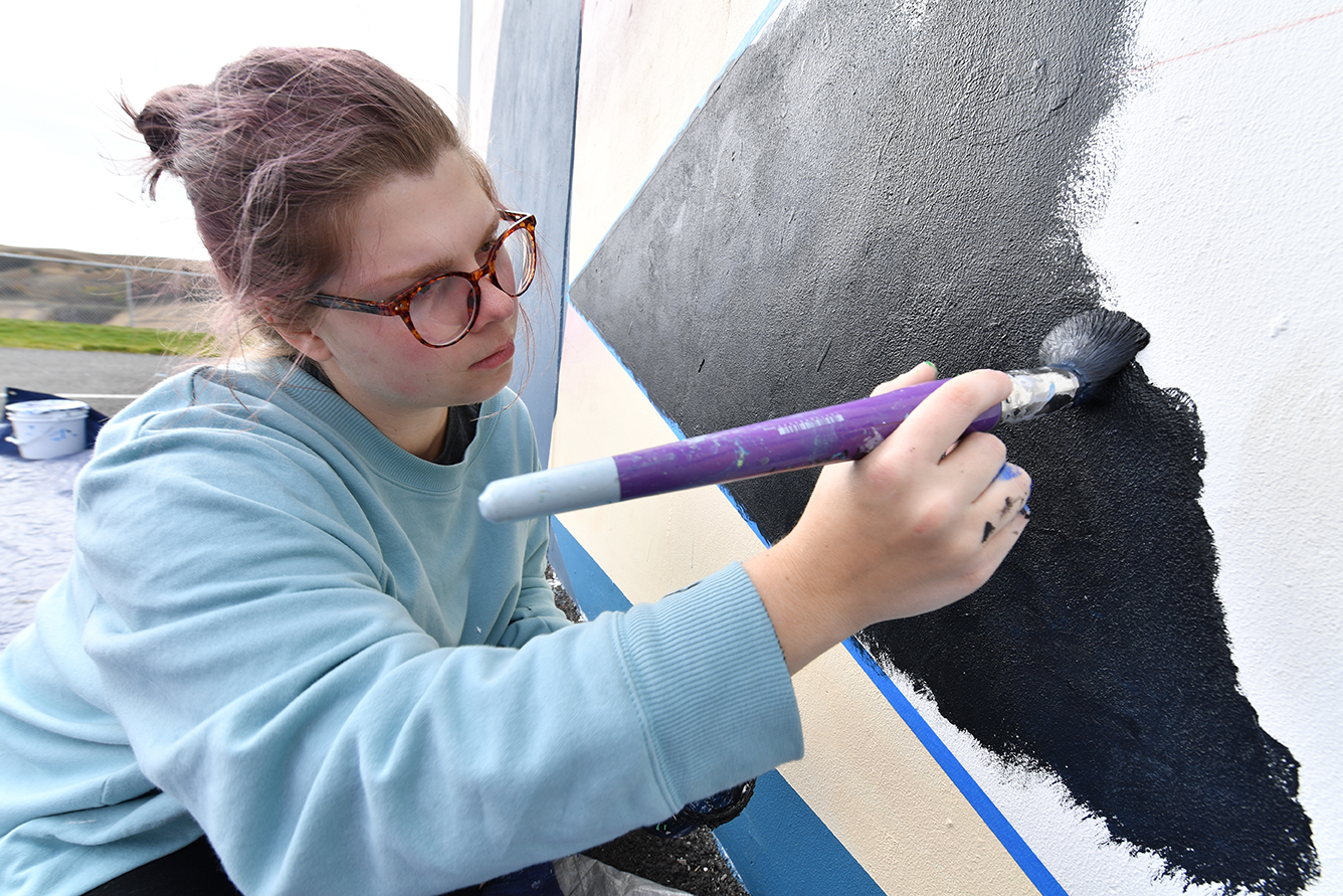 An artist wearing glasses painting with a brush on a large dark canvas