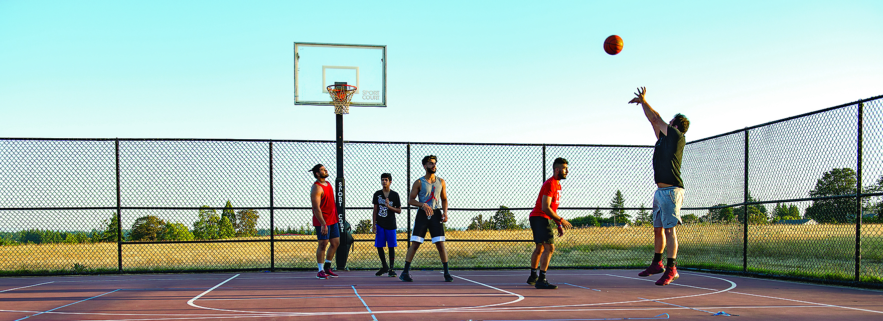 Young adults playing basketball on an outdoor court under a clear blue sky