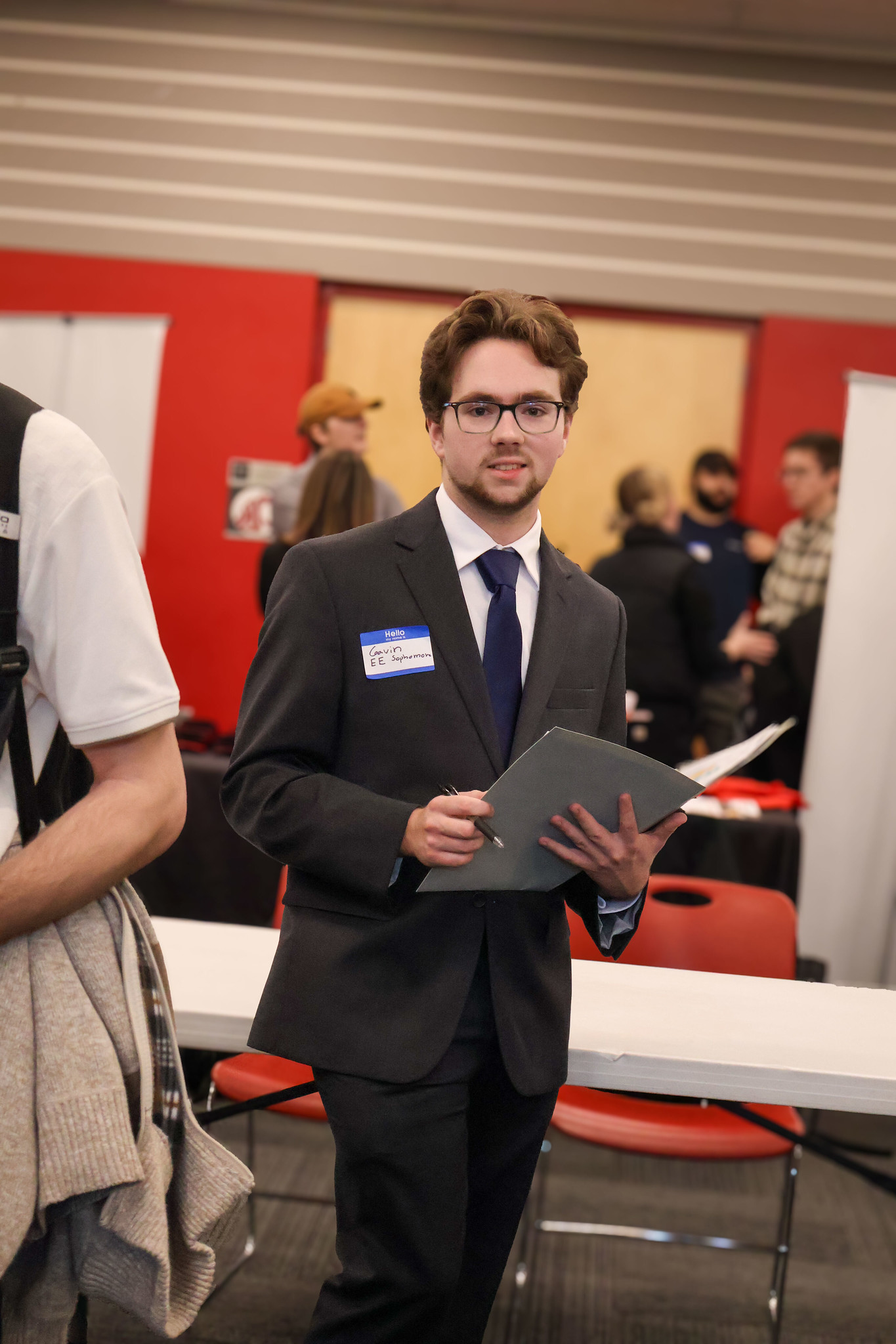 A man in a suit jacket holding a folder and speaking at a corporate or academic event