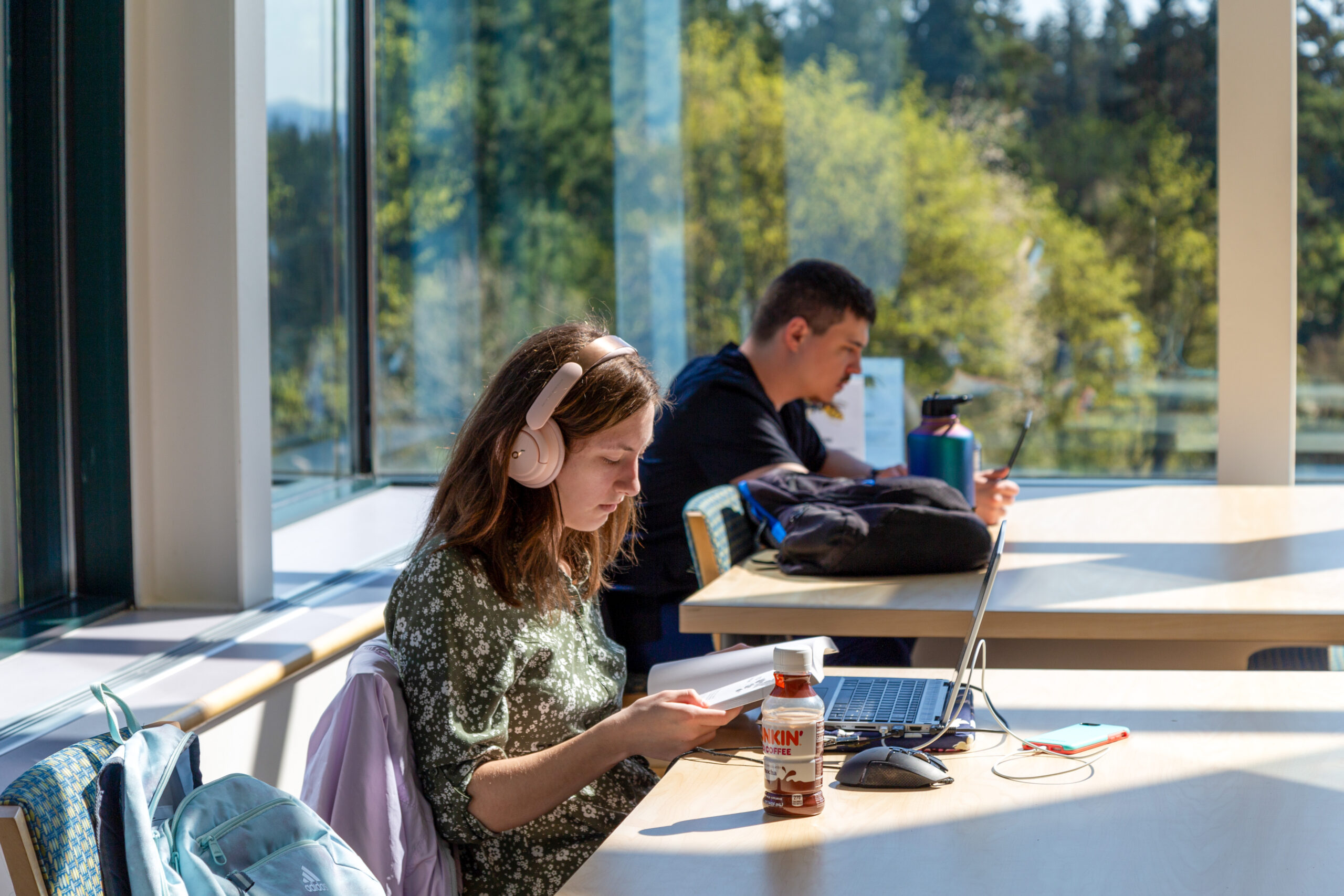 Students sitting at a high-top table working on laptops by a large window