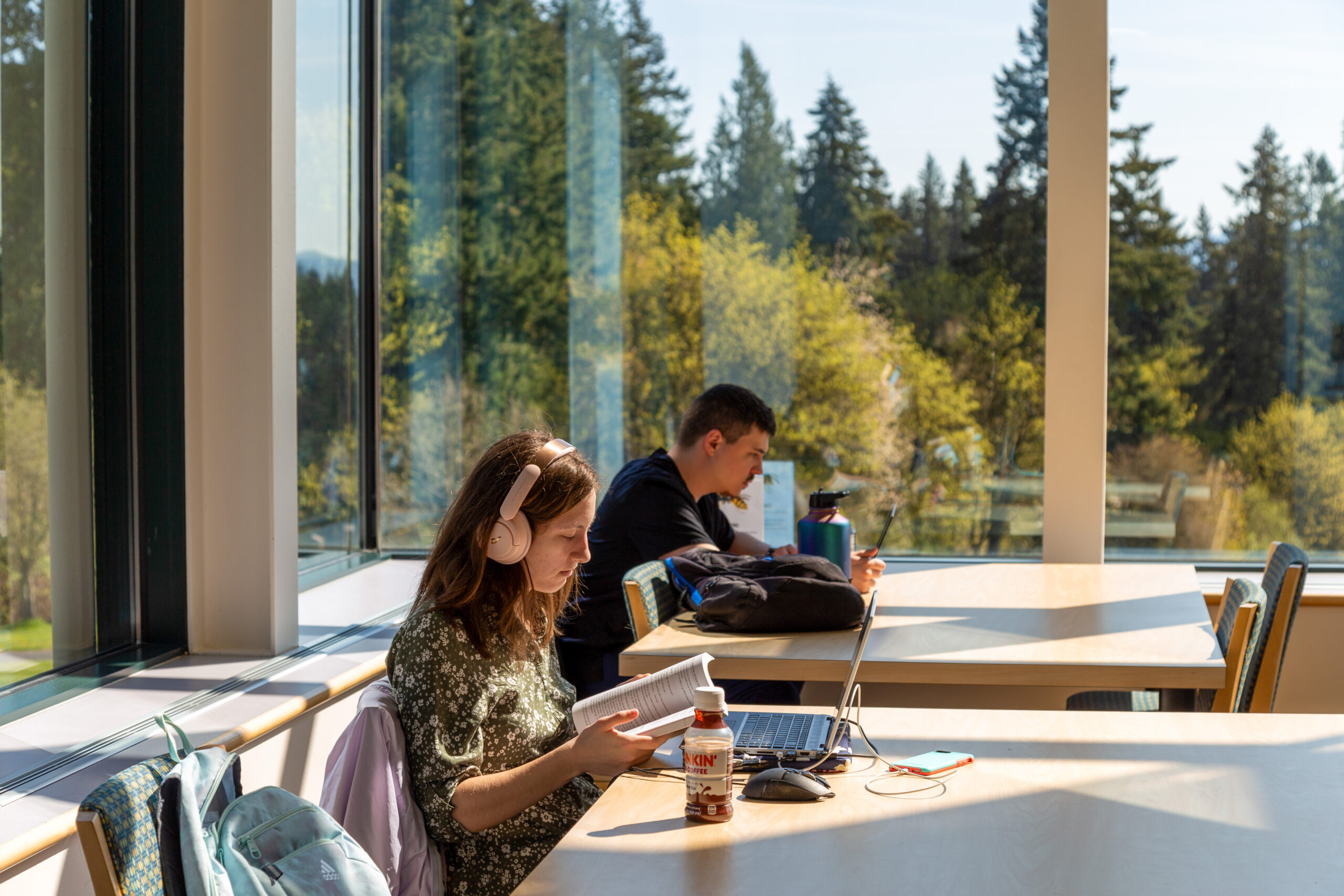 A female student wearing headphones reads in the library, with trees in the background