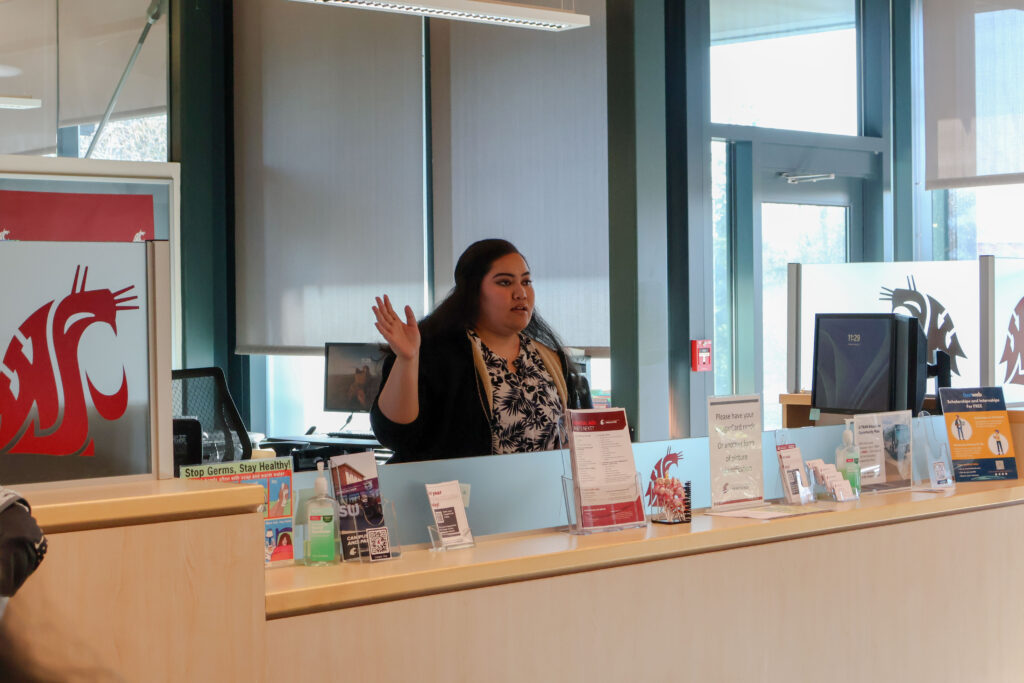 A presenter speaking to an audience from behind a reception desk at an indoor event