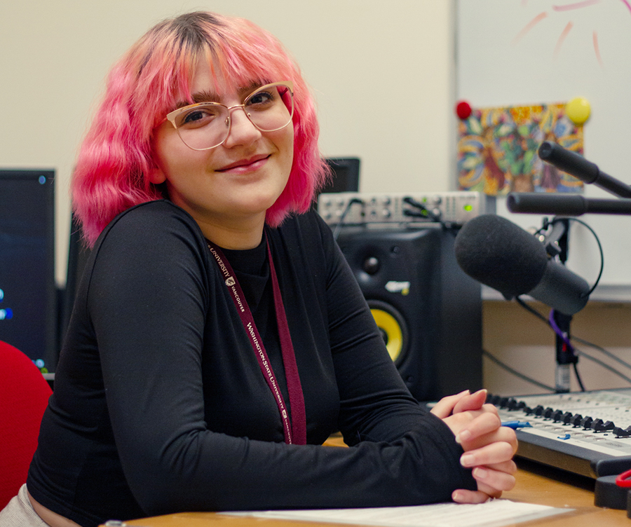 Portrait of a smiling student with pink hair sitting at an indoor radio broadcasting desk on campus
