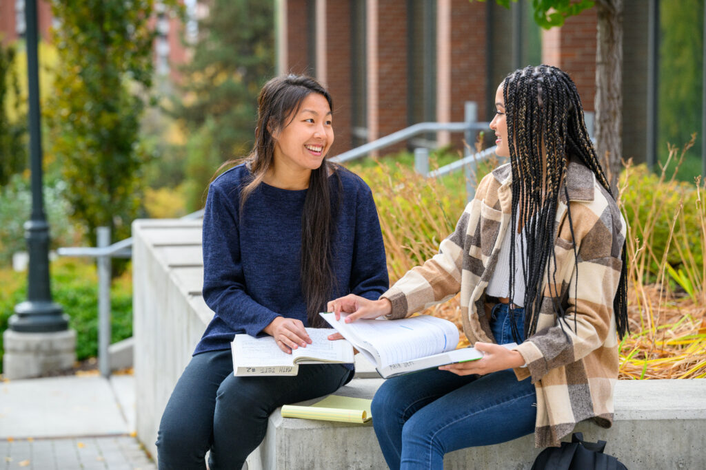 Two female students sitting outside on campus steps reviewing course materials together