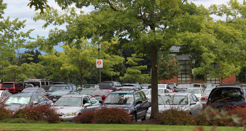 A full parking lot at WSU Vancouver viewed through the shade of green trees and bushes.