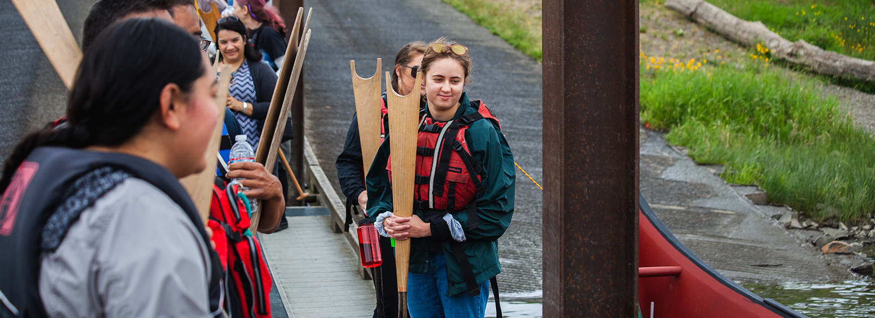 A student standing next to a campus pathway holding large wooden boards during an outdoor activity