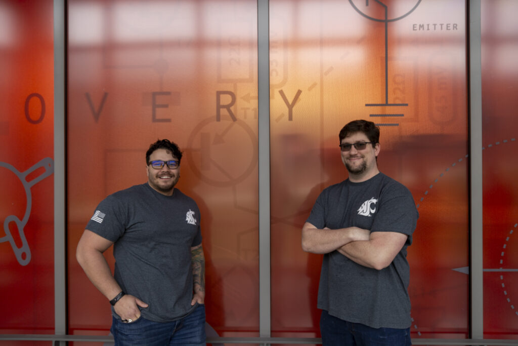 Veteran and Military-Affiliated Student Services - WSU Vancouver - 2 Veterans standing infront of sign | Two student veterans wearing matching group shirts standing and posing together outdoors on campus