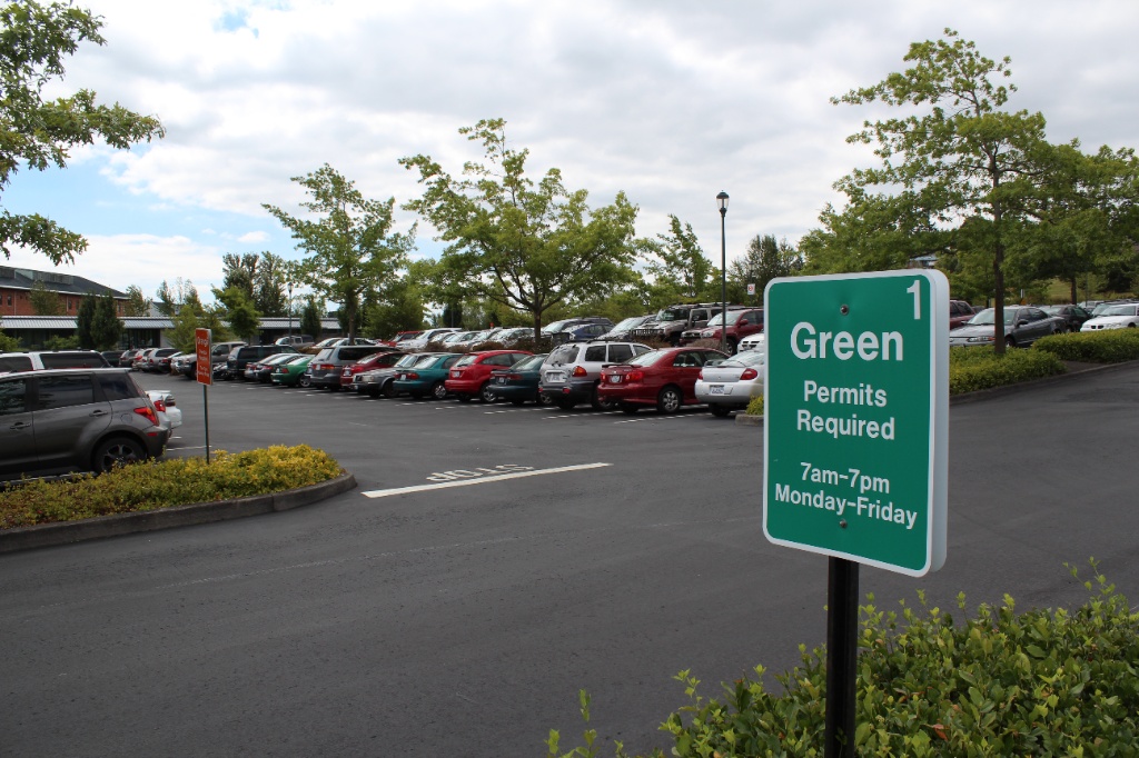 A green 'Green Permits Required' sign stands at the entrance of an asphalt campus parking lot packed with cars.