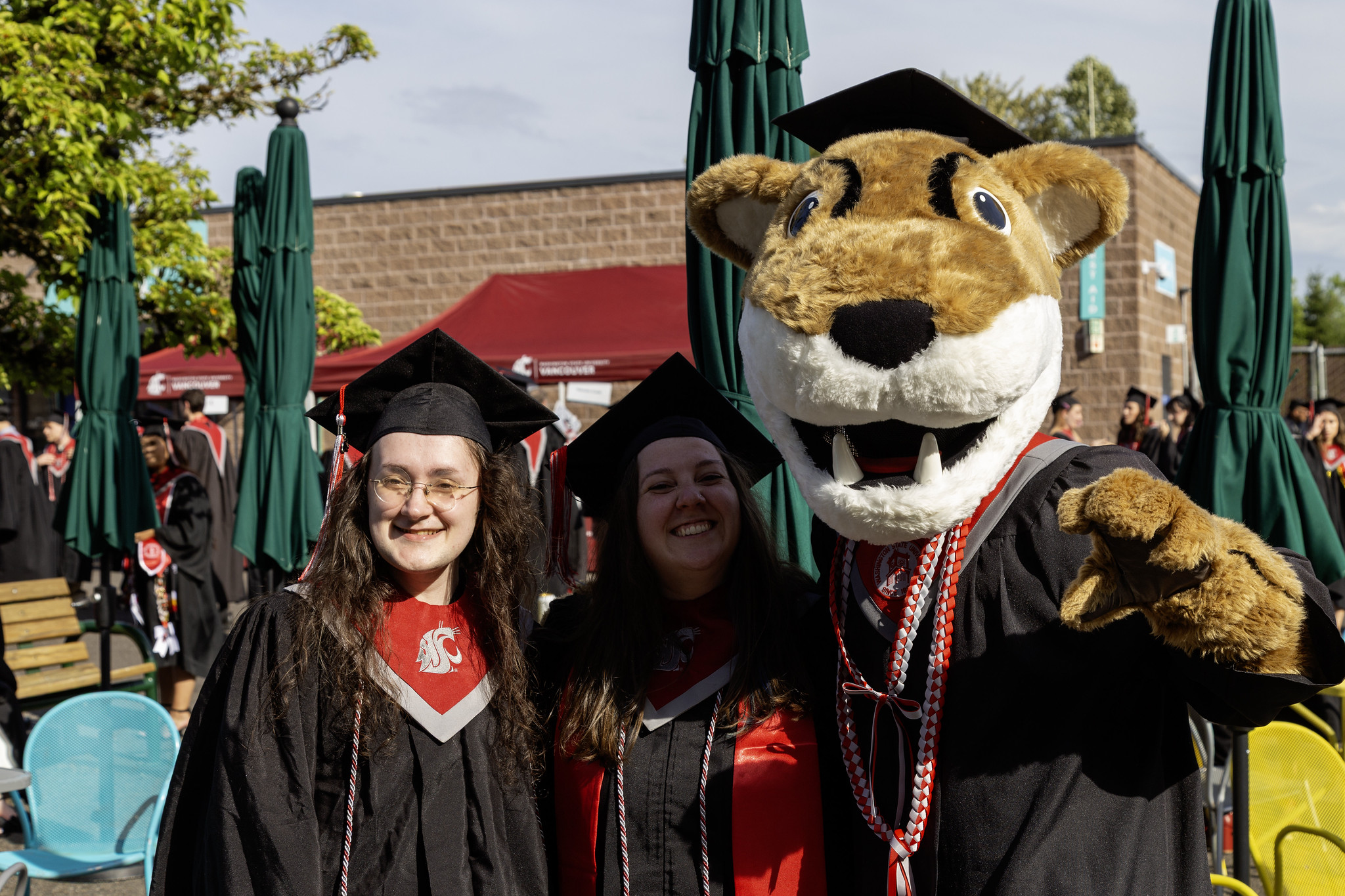 Two smiling graduates in black caps and gowns stand with the WSU mascot, Butch Cougar, at an outdoor WSU Vancouver commencement celebration.