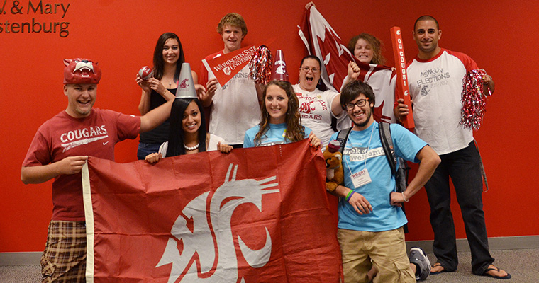 A group of cheerful students holding up large crimson flags at a WSU Vancouver outdoor event