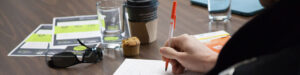 A close-up of a person's hand writing with a pen on a paper notepad at a desk