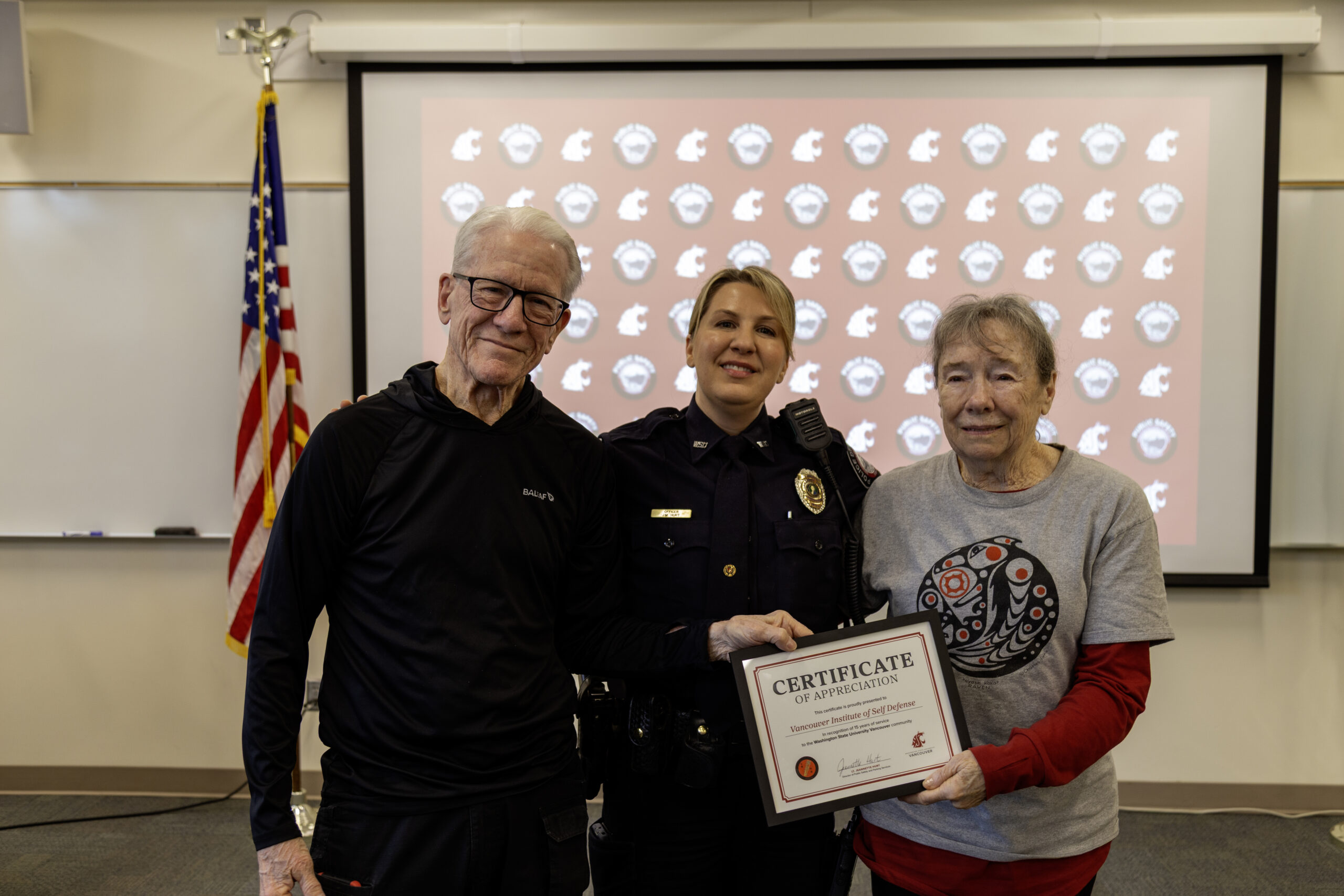 A police officer stands with two community members holding a completion certificate in a classroom.