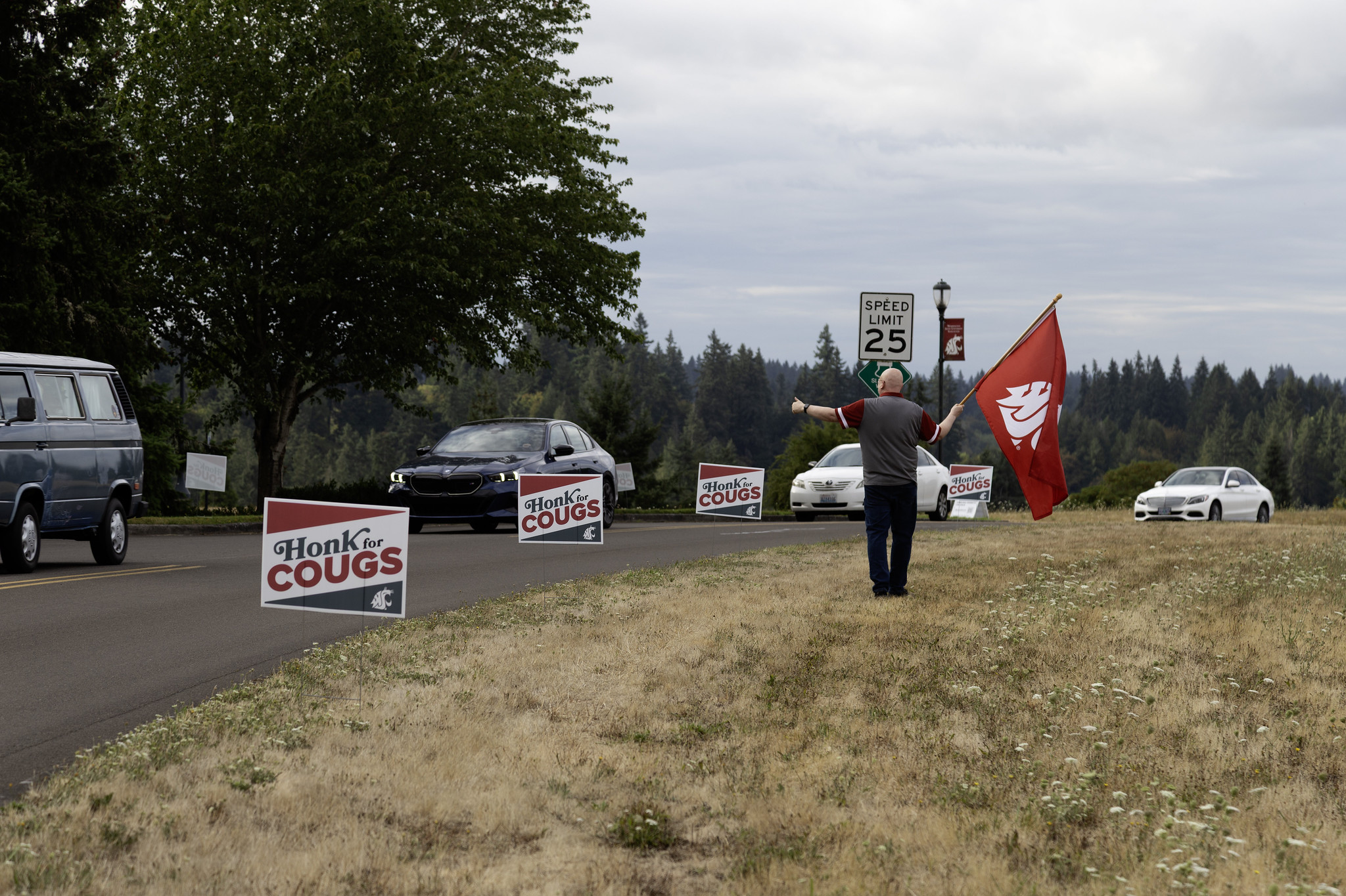 A roadside view at WSU Vancouver featuring cars, 'Honk for Cougs' yard signs, a person holding a large red WSU flag, and a background of evergreen trees under an overcast sky.