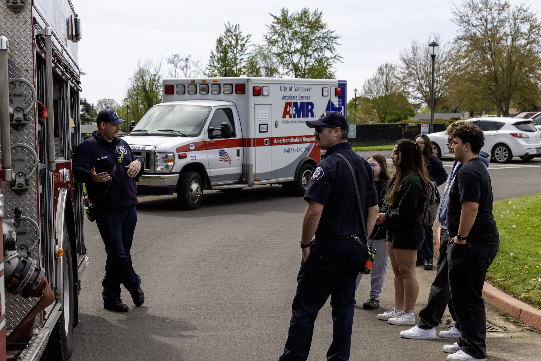 An AMR ambulance and a fire engine parked outdoors on campus with emergency responders and a group of students gathered nearby.