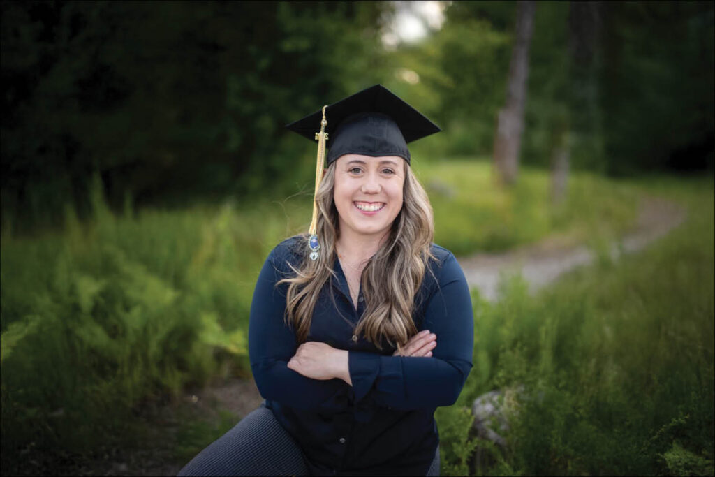 A student in a graduation cap and gown sitting smiling in a green field