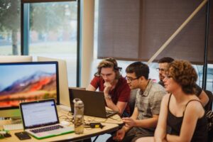Students sitting in a row working on laptops at a long table by a window