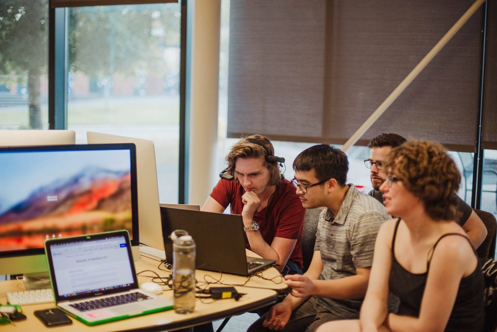 Students sitting in a row working on laptops at a long table by a window