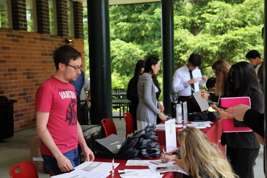 A group of people gathered around an outdoor registration table at a WSU Vancouver event, with a staff member in a red T-shirt assisting attendees.
