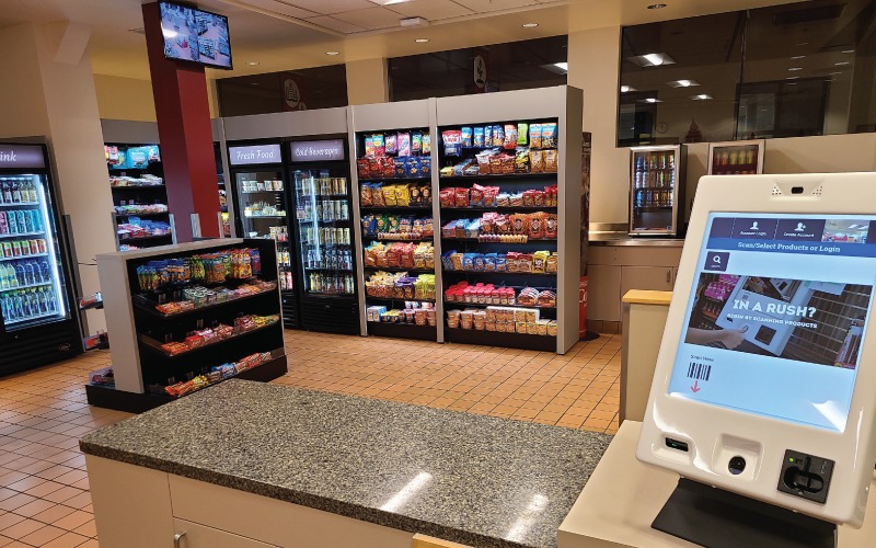 An interior view of the self-service micro-market on the WSU Vancouver campus, featuring snack shelves, beverage coolers, and an automated self-checkout kiosk terminal in the foreground.