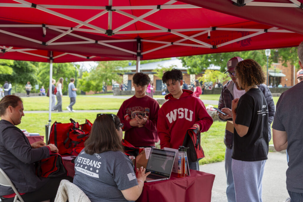 A group of students gathering around an outdoor campus information table under a red canopy tent, where a university staff member wearing a WSU Vancouver Veterans shirt assists them on a laptop.