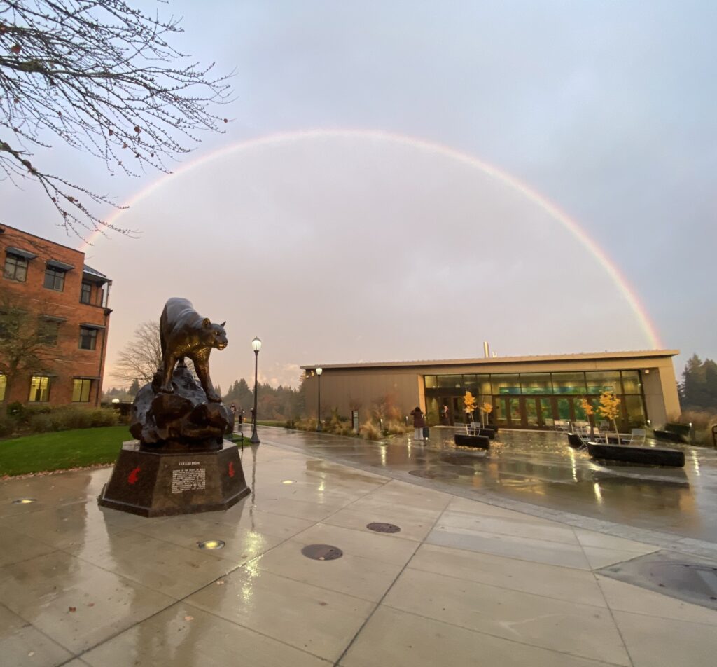 A wide, vibrant rainbow spanning across the sky over the WSU Vancouver campus plaza and the Firstenburg Student Commons on a rainy day.