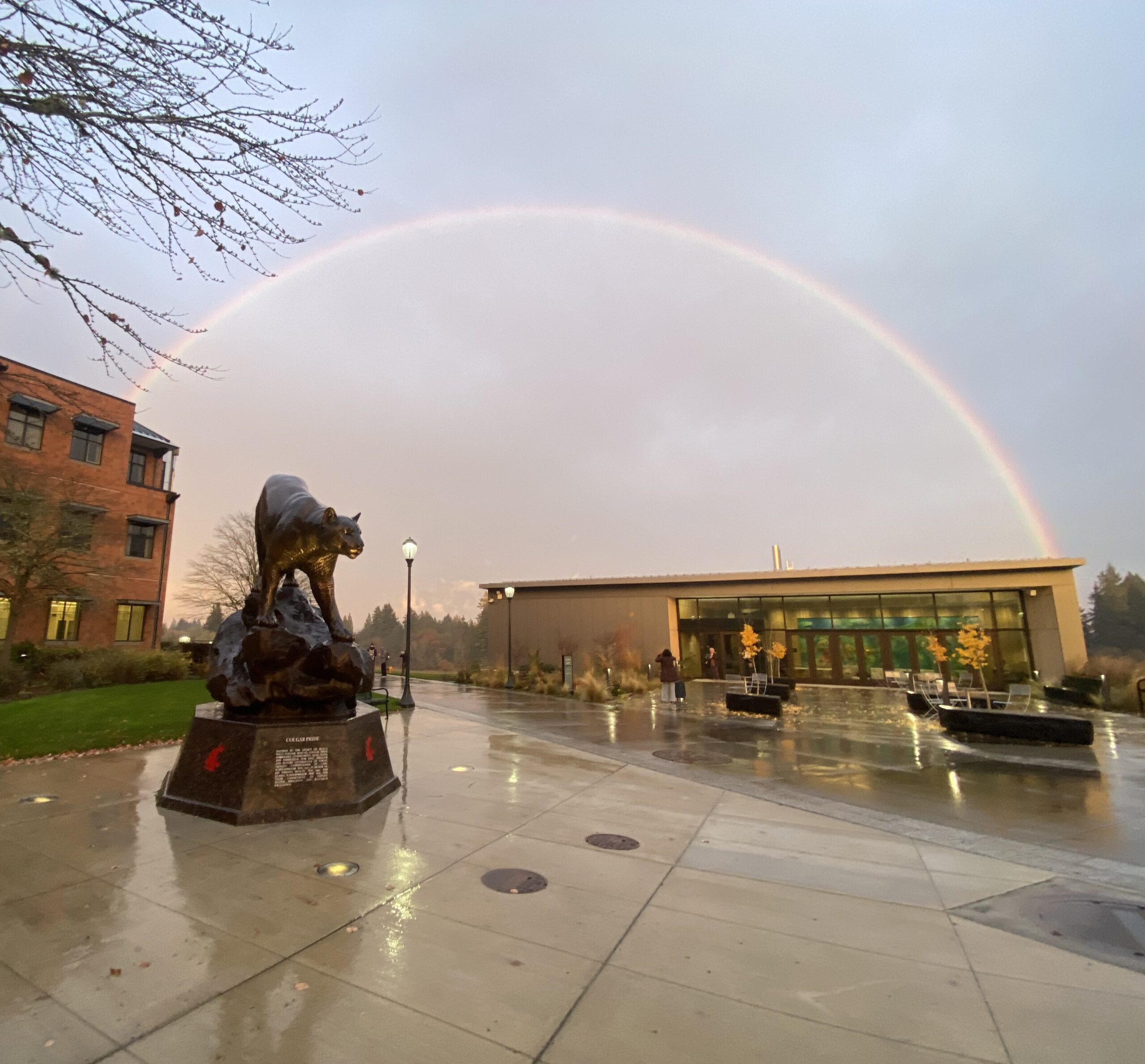 A wide, vibrant rainbow spanning across the sky over the WSU Vancouver campus plaza and the Firstenburg Student Commons on a rainy day.