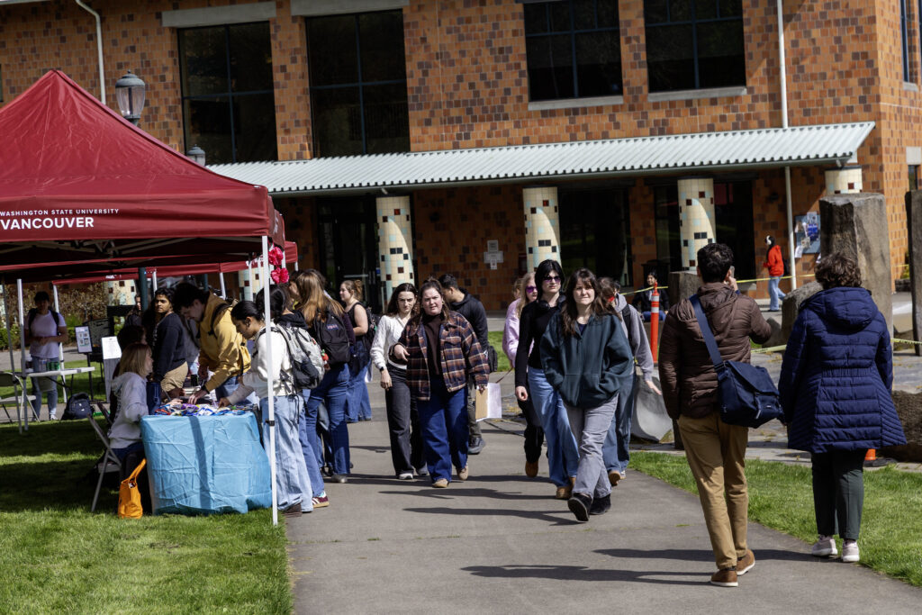 Students walking on a campus pathway past outdoor club information tables outside a brick building at WSU Vancouver.