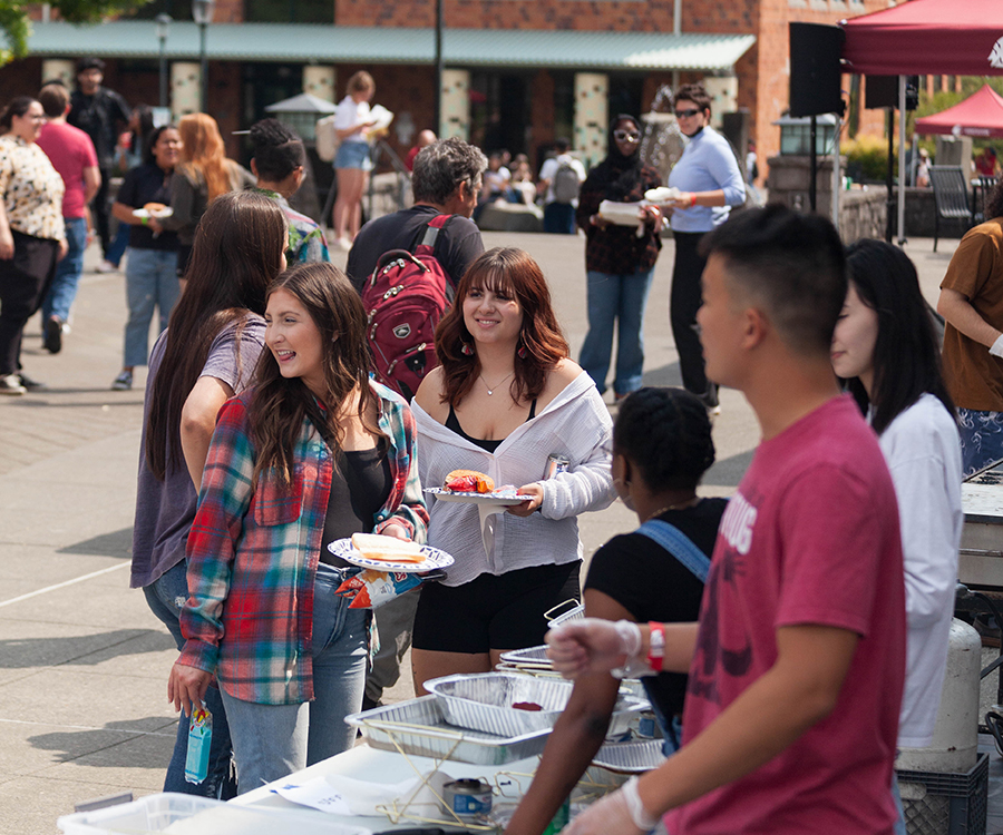 Students gather on the campus quad for the Back to School Barbecue as volunteers serve food from metal trays outside on a sunny day.