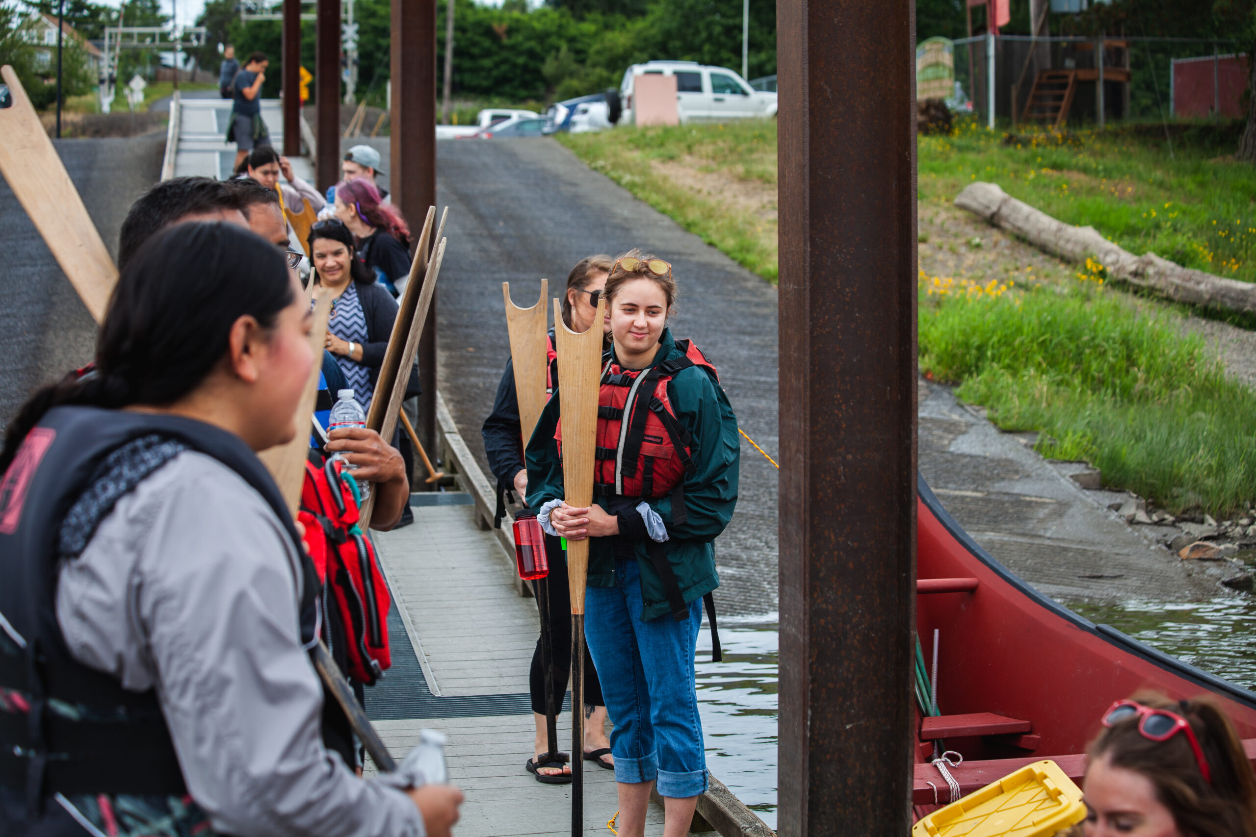 A group of students wearing life vests and holding wooden oars gather on a loading dock before boarding canoes for a lake excursion.