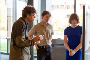 Three attendees review and discuss a research poster near floor-to-ceiling windows during a poster presentation session.