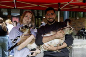 Two smiling WSU Vancouver students holding baby goats under a red canopy tent outdoors.