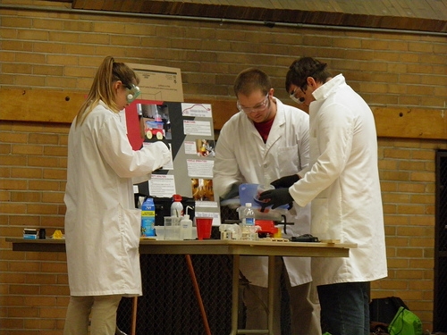 Students in lab coats and safety glasses working on a table.