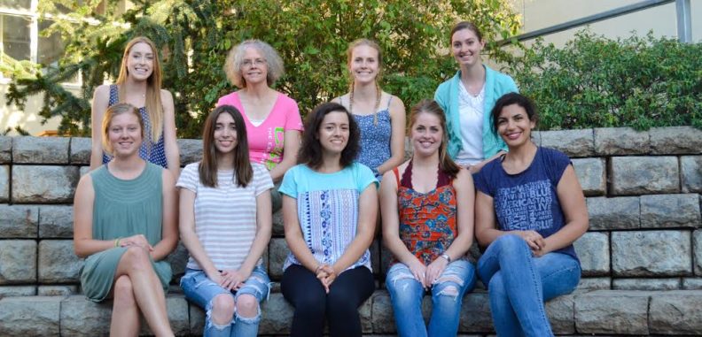 Nine women seated in two rows in front of a block wall.