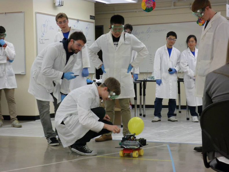 Students wearing safety gear looking at a chemical car that incorporates a yellow balloon.