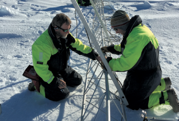 Two men kneeling in the snow near atmospheric equipment.