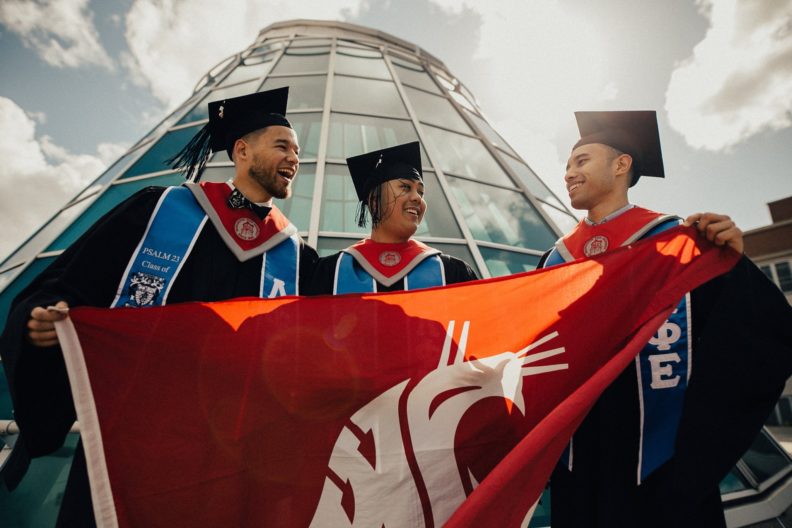 Three WSU students wearing graduation caps and gowns and holding a WSU Cougar flag.