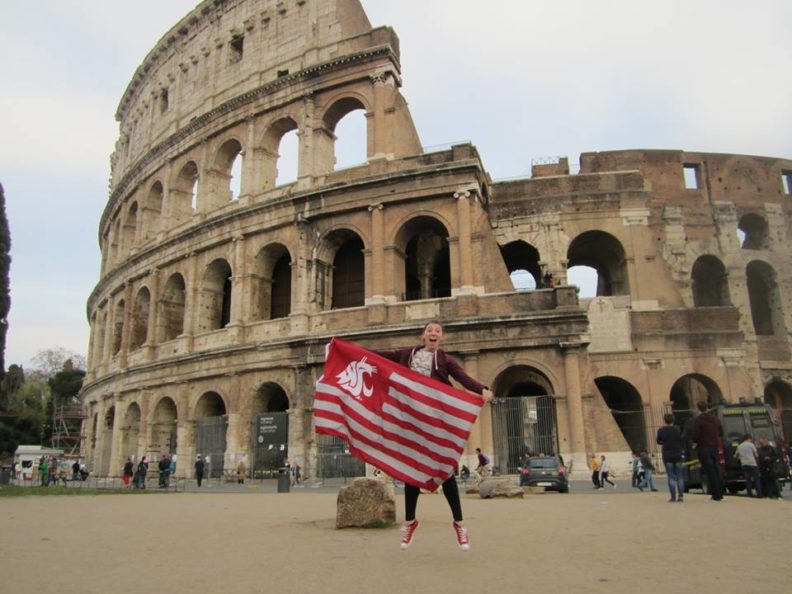 Madison Newberry mid jump in front of the Roman Colosseum while holding a WSU Cougar flag.