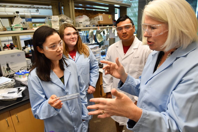 Mary Rezac and three students in a lab wearing lab coats and safety glasses.