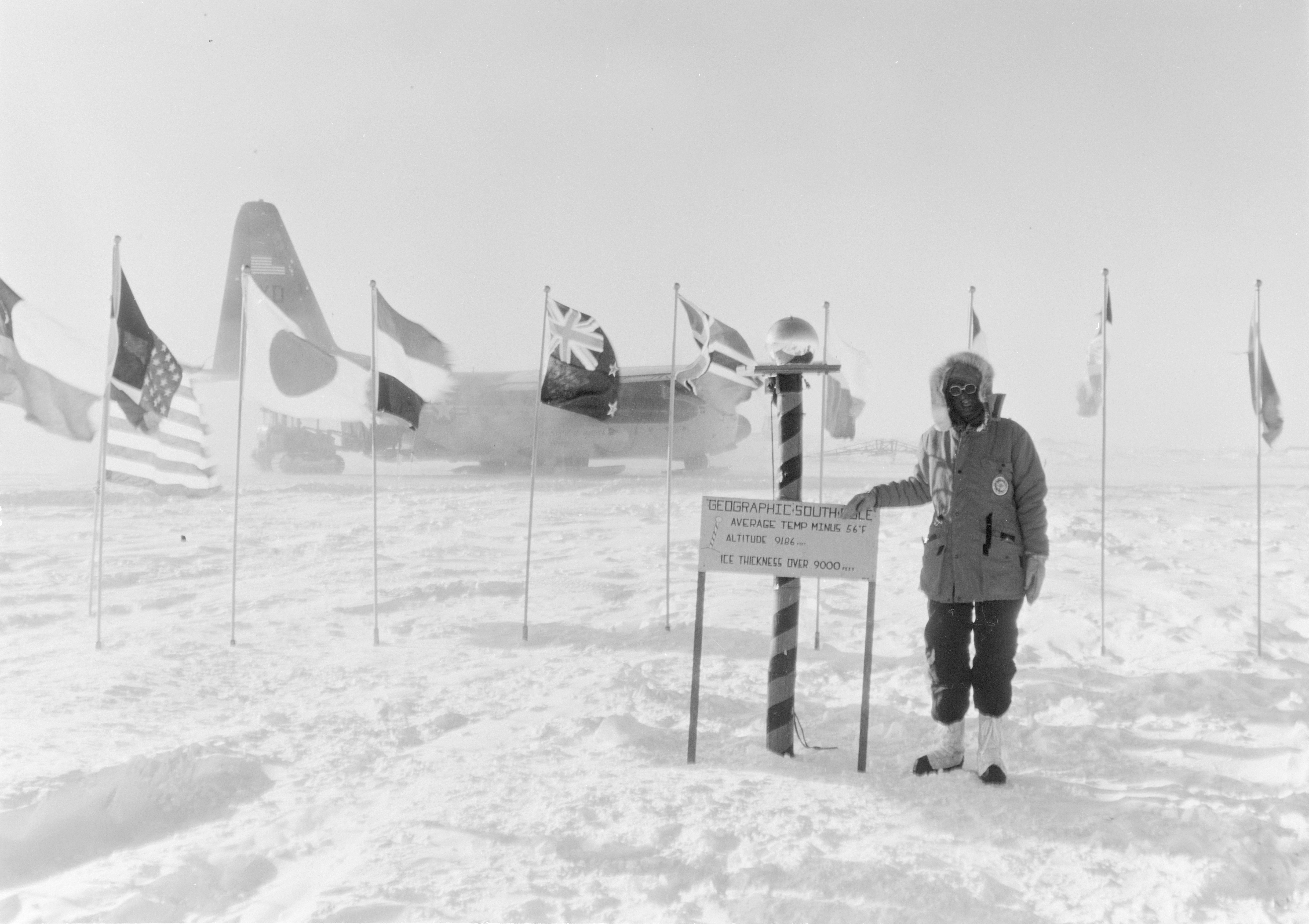 Person next to a sign that reads Geographic South Pole, with international flags and a plane in the background.