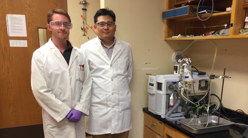 Jake Gray and Su Ha standing in a lab wearing white lab coats.
