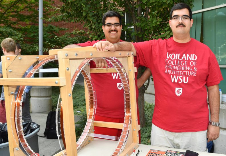 Joseph and Mario Guerrero stand next to a square wood structure housing a metal wheel.