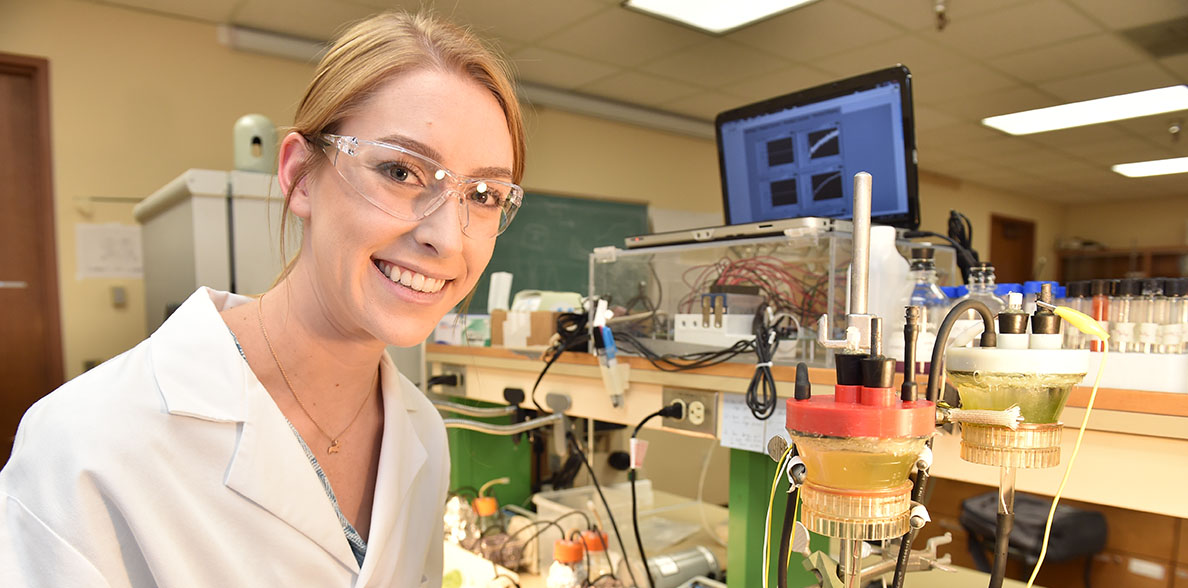 Roslyn VanSickle seated at a lab station wearing a lab coat and safety glasses.